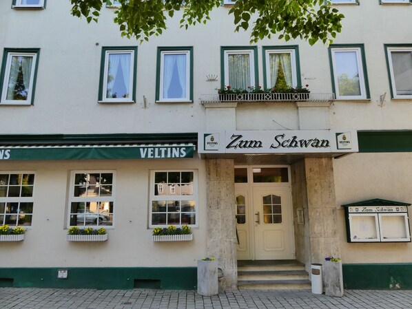 A building with a sign reading "Zum Schwab" and "VELTINS" above the entrance. There are flower boxes on the windows and a small balcony with plants.