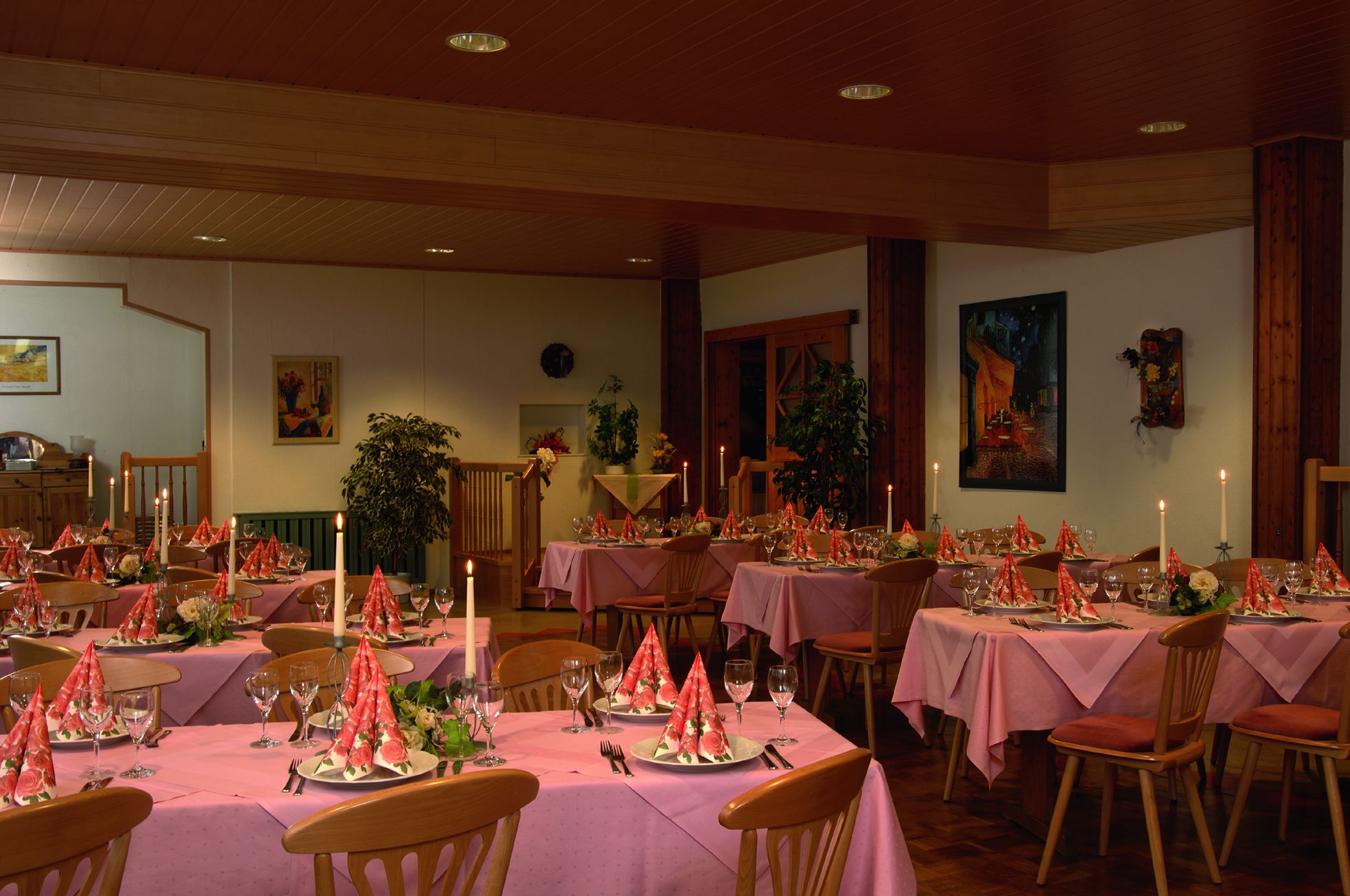 Un comedor con varias mesas dispuestas para una comida, con manteles rosas y centros de mesa florales rojos.
