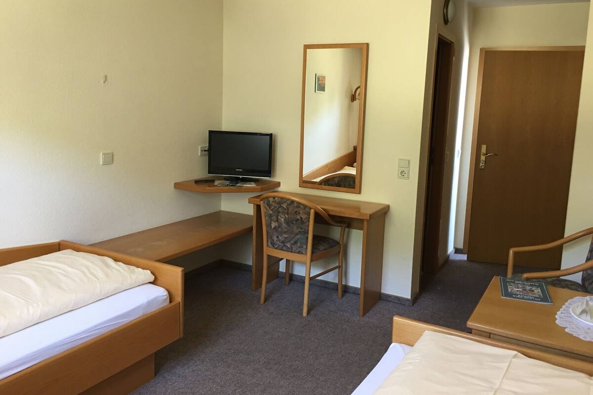 A hotel room with a bed, a desk with a computer, a chair, a mirror, and a door.