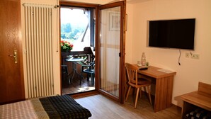 A room with a bed, a desk, a chair, a TV, and a sliding glass door leading to a balcony with a table and chairs. - Blick zum Maimont (Ludwigswinkel)