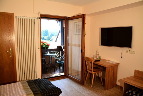 A room with a bed, a desk, a chair, a TV, and a sliding glass door leading to a balcony with a table and chairs.