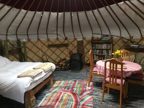 Un interior acogedor de una yurta con una cama, una silla de madera, una mesita con mantel a cuadros, una estantería y una alfombra colorida.