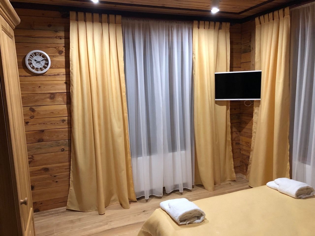 A room with wooden panel walls, a bed with a white bedspread, two towels, a wall clock, and a TV mounted on the wall.