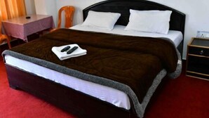 A single bed with a dark headboard, white pillows, and a brown blanket. There is a small table with a chair beside the bed, and a nightstand with a lamp. - Hotel Taj Himalaya khirsu (Srinagar)