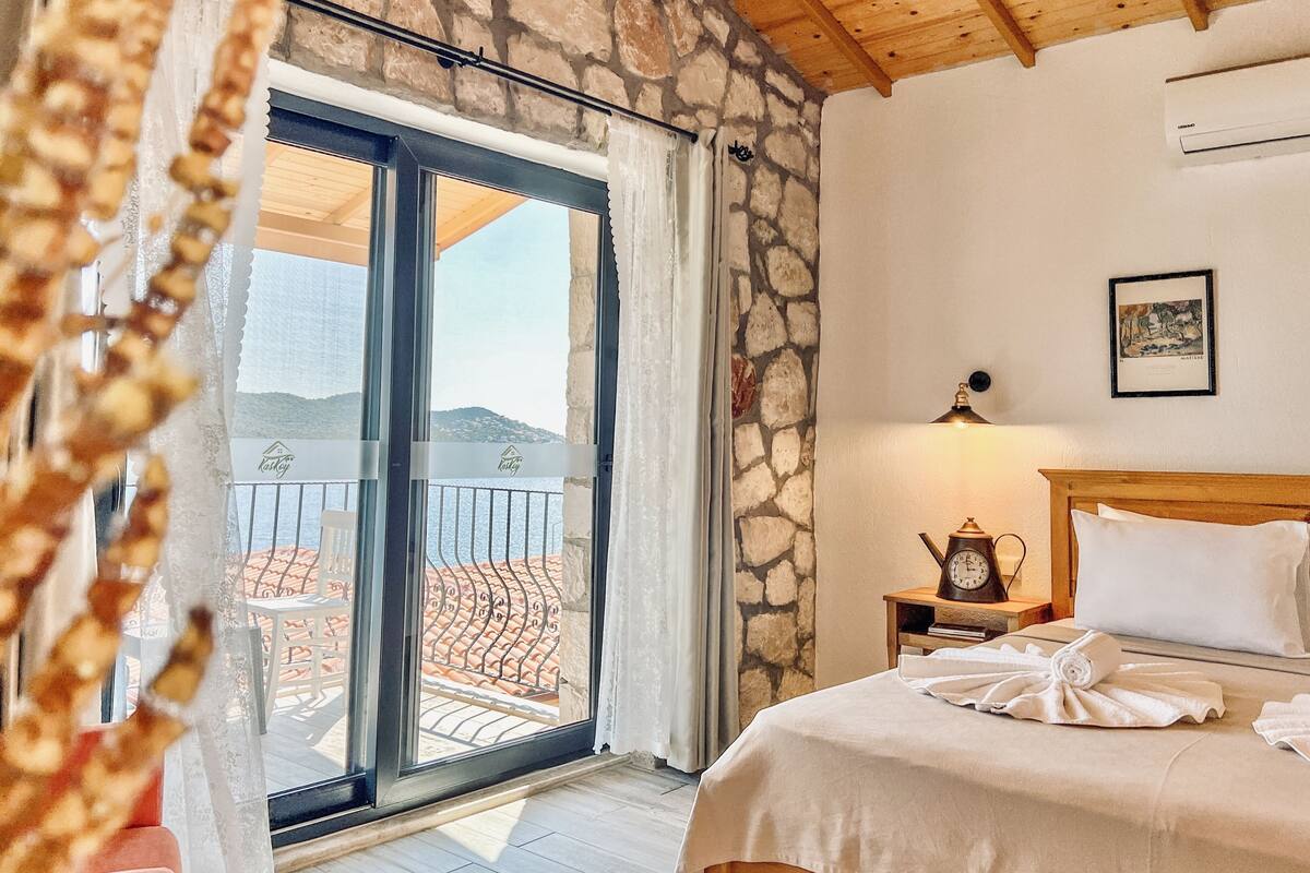 A hotel room with a bed, a nightstand with a clock, a framed picture on the wall, and a balcony with a view of the sea.