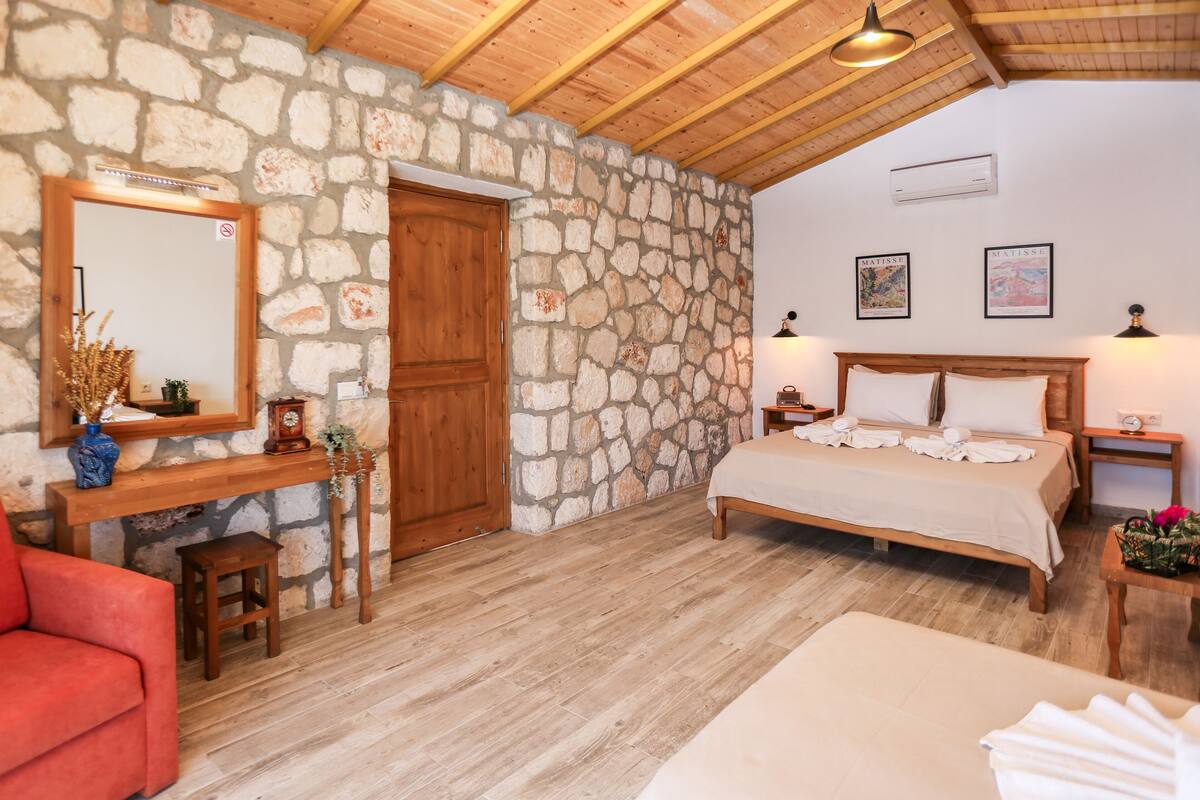 A bedroom with a stone wall, wooden ceiling, a bed, a nightstand, a desk, a chair, and a mirror.