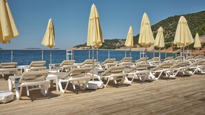 A beachside area with rows of white lounge chairs and yellow umbrellas, overlooking a clear blue sea and a hilly landscape in the background. - Kaskoy Hotel (Kas)