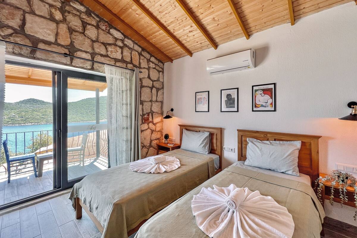A room with two beds, a stone wall, wooden ceiling beams, an air conditioning unit, and a balcony with a view of the sea.