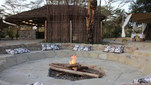 A stone fire pit with logs burning, surrounded by stone seating with patterned cushions. - Swara Acacia Lodge (Athi River)