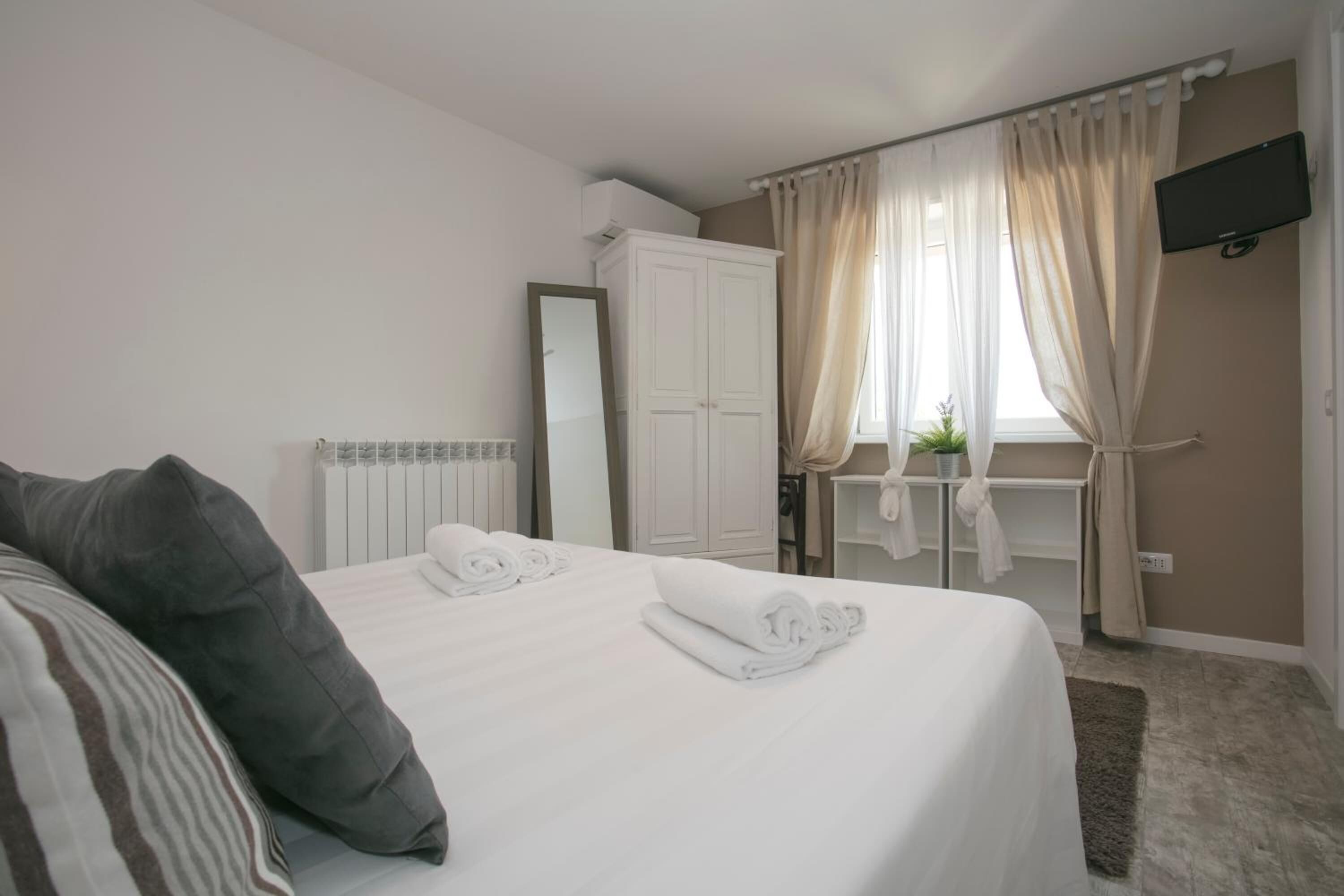 A bedroom with a bed, a mirror, a wardrobe, a window with curtains, and a television on the wall.