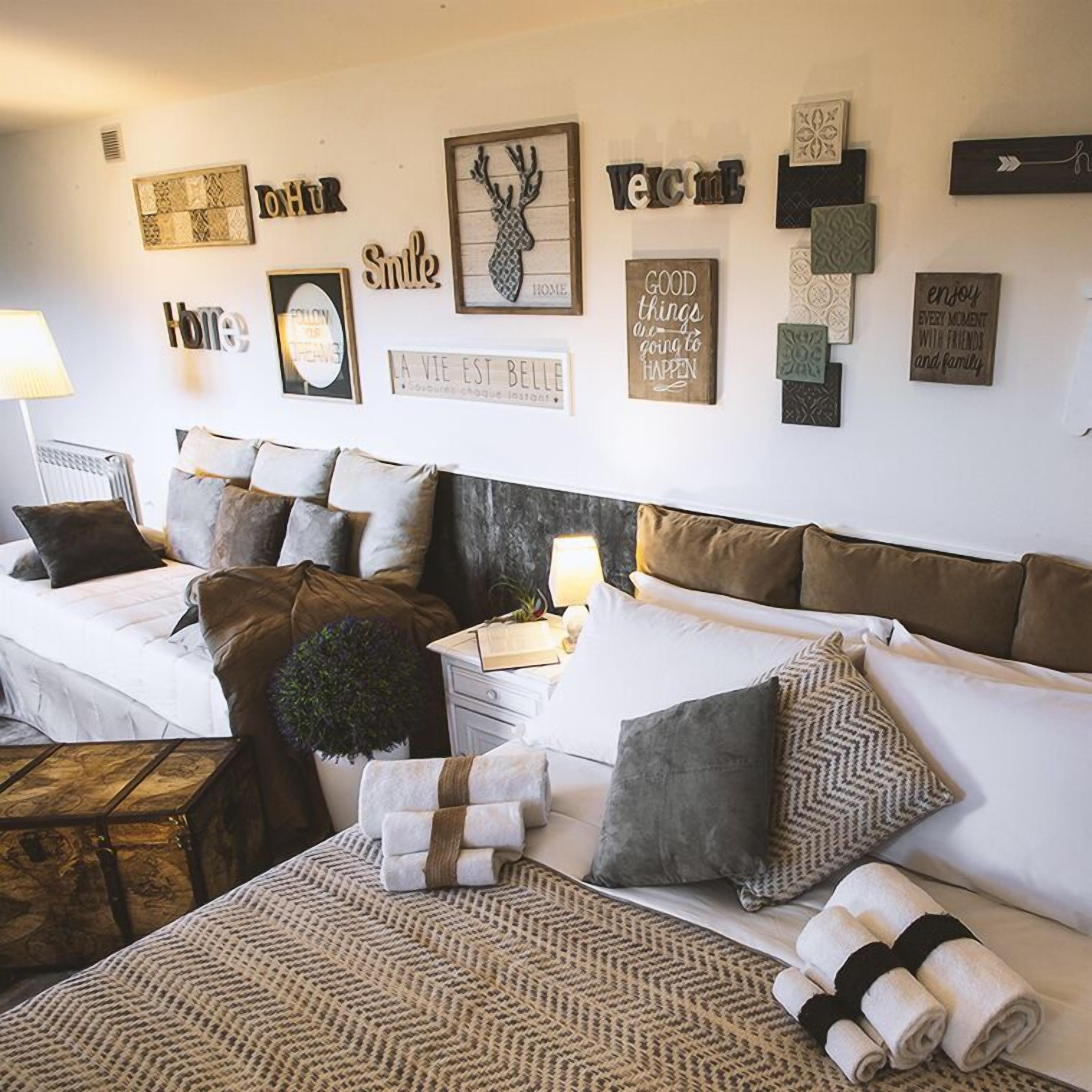 A cozy bedroom with a bed, a nightstand, and a bedside table. There are multiple pillows and a blanket on the bed. The wall has various decorative items and signs.
