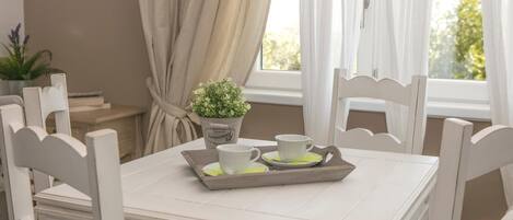 Une table à manger avec deux chaises blanches, un plateau avec deux tasses et soucoupes blanches, une plante en pot et une fenêtre avec des rideaux.