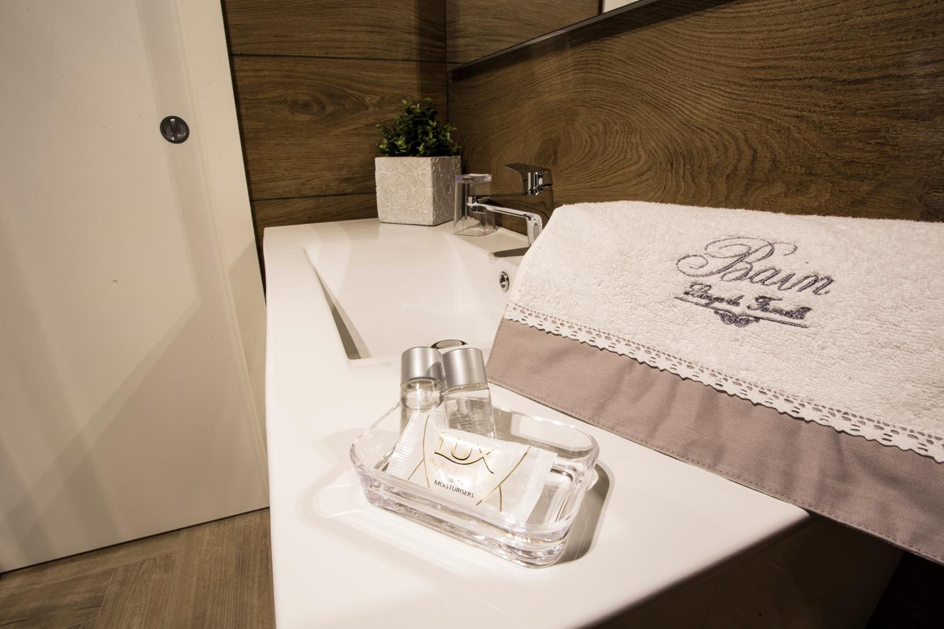 A bathroom with a white sink, a clear bottle of lotion, and a towel with "Baum" embroidered on it.