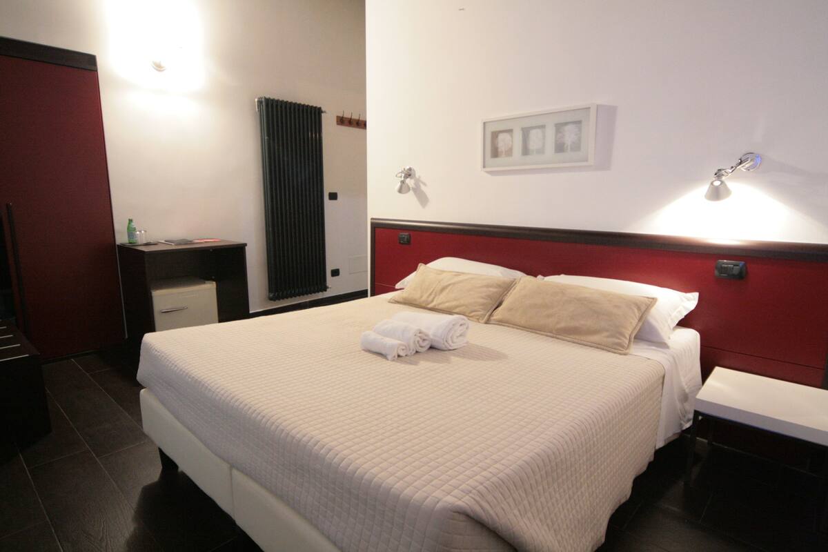 A neatly made bed with white bedding and two pillows, a bedside table with a lamp, and a red accent wall.