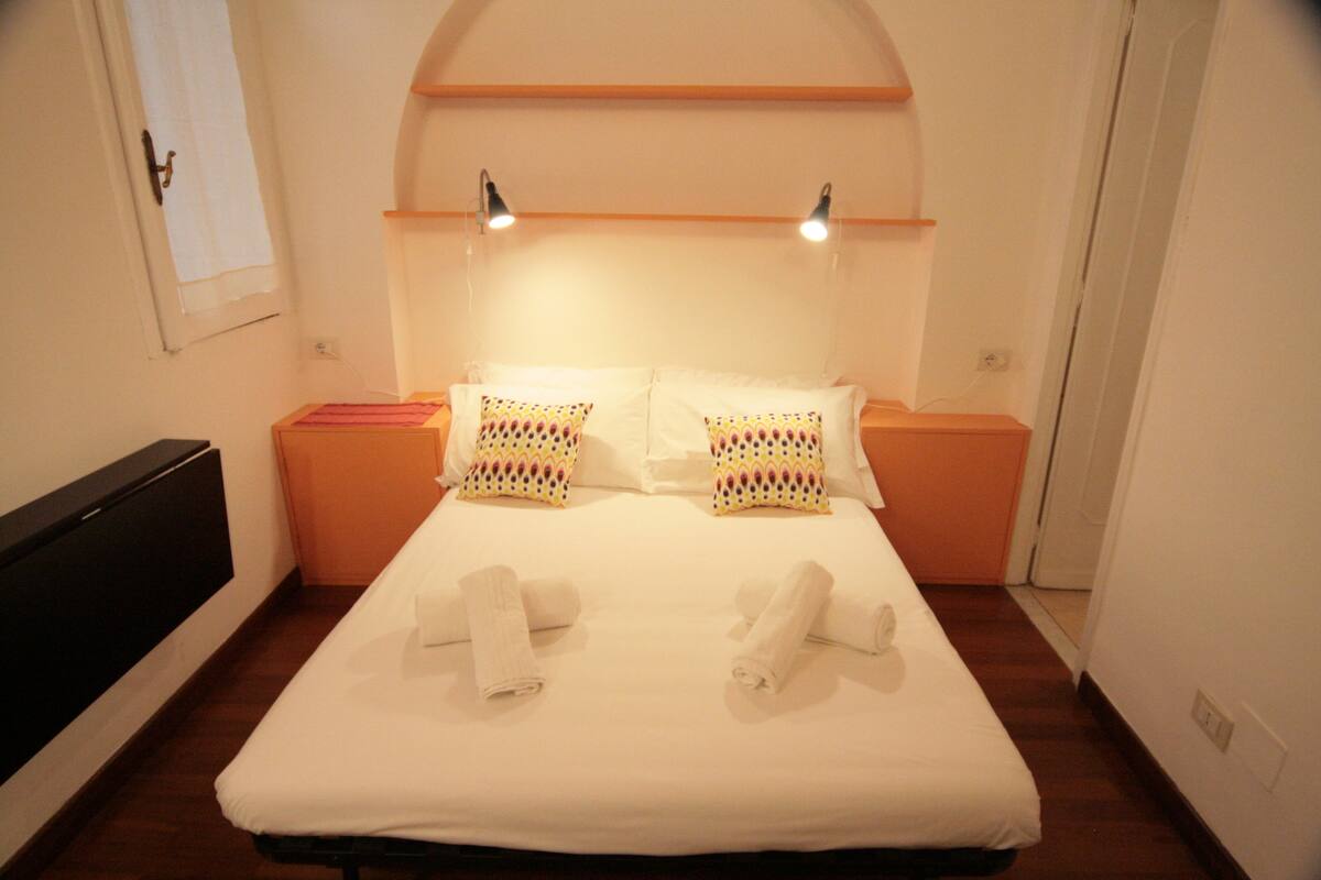 A neatly made bed with white linens and two pillows, a bedside table with a pink object, and a wall-mounted shelf with two lamps.