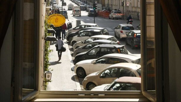 Une vue sur la rue, prise depuis une fenêtre avec des rideaux, montrant des voitures garées, des immeubles et un piéton.