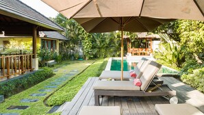 Zona junto a la piscina con sillones de descanso, terraza de madera y una gran sombrilla que da sombra.