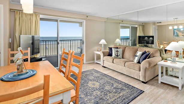 A living room with a dining area, a sofa, a TV, and a balcony with a view of the sea.