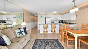 A compact living space with a kitchenette, a dining area with wooden chairs, a sofa with patterned cushions, and a blue rug.