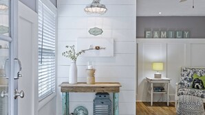 Een hal met een witte deur, een houten console en decoratieve objecten, een bordje aan de muur, en een zithoek met een lamp en gekleurde kussens.