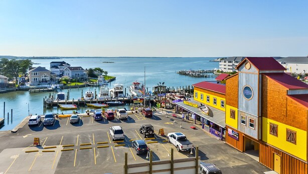A marina with boats docked, a parking lot with cars, and buildings with a red roof and a yellow facade. - Stylish, upscale luxury 4-bedroom condo with free WiFi and an outdoor pool located downtown on the bay water and just a few blocks to the beach! (Ocean City)