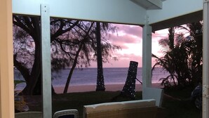 Un espace extérieur aménagé avec des meubles en osier, abrité et offrant une vue sur une plage bordée d’arbres et un magnifique coucher de soleil.