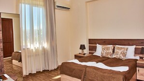 Una habitación de hotel con una cama grande, cabecera de madera, mesita de noche con lámpara y ventana con cortinas traslúcidas.
