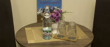 Ett runt träbord med en flaska Borjomi-mineralvatten, två glas och en vas med lila blommor. En affisch med en bild av Georgien hänger på väggen i bakgrunden.