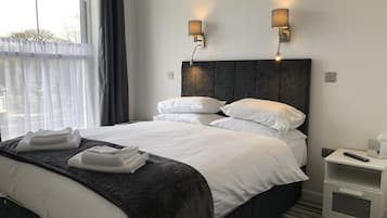 En pænt redt seng med hvide sengetøj og en mørk sengegavl, to vægmonterede lamper, et natbord med en fjernbetjening og et vindue med lette gardiner.