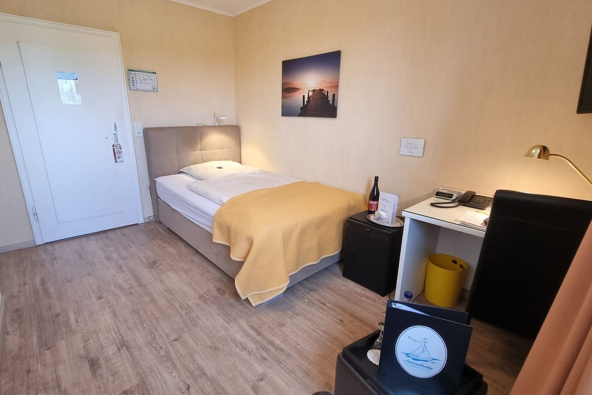 A hotel room with a bed, bedside table, lamp, and a small desk with a telephone and a trash bin.