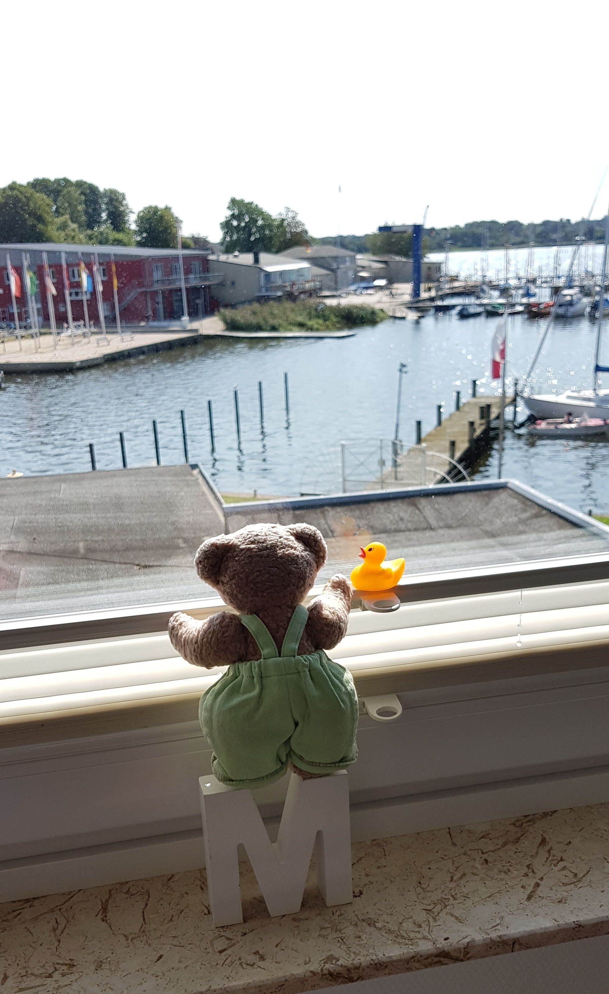 Un oso de peluche sobre un taburete con forma de letra 'M' junto a una ventana que da a un puerto deportivo con barcos.
