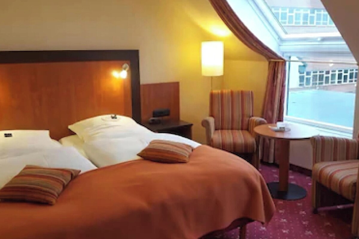A hotel room with two beds, a small table, two armchairs, a lamp, and a view of the outside.