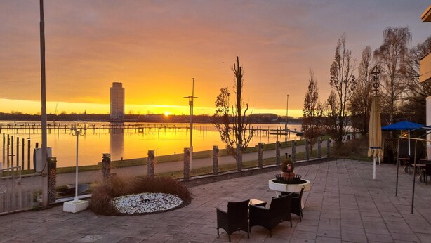 Een buitenpatio met uitzicht op een zonsondergang boven het water, met een hoog gebouw, een steiger en een kleine tuin met stoelen.