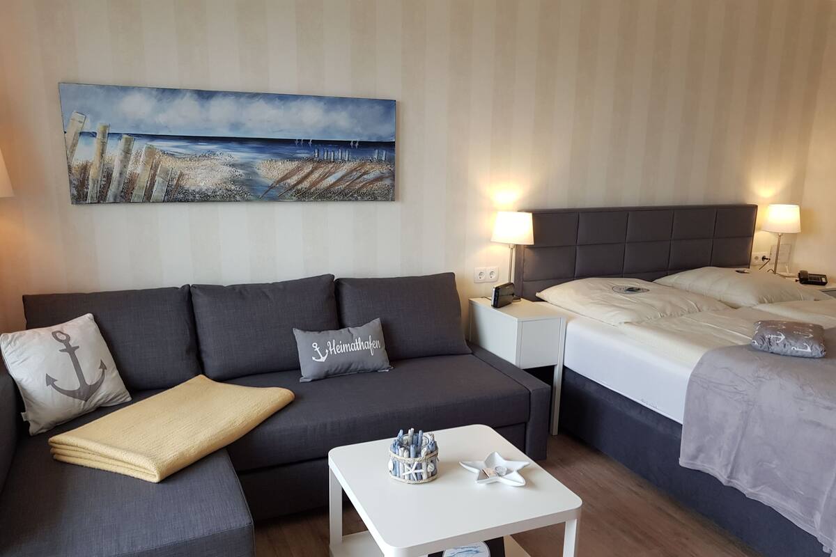 A modern hotel room with a grey sofa, a bed, a nightstand, a bedside table, and a painting of a beach scene.