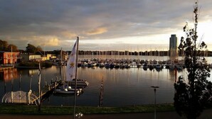 Een jachthaven met aangemeerde boten, een zeilboot met vlag en een reuzenrad op de achtergrond.