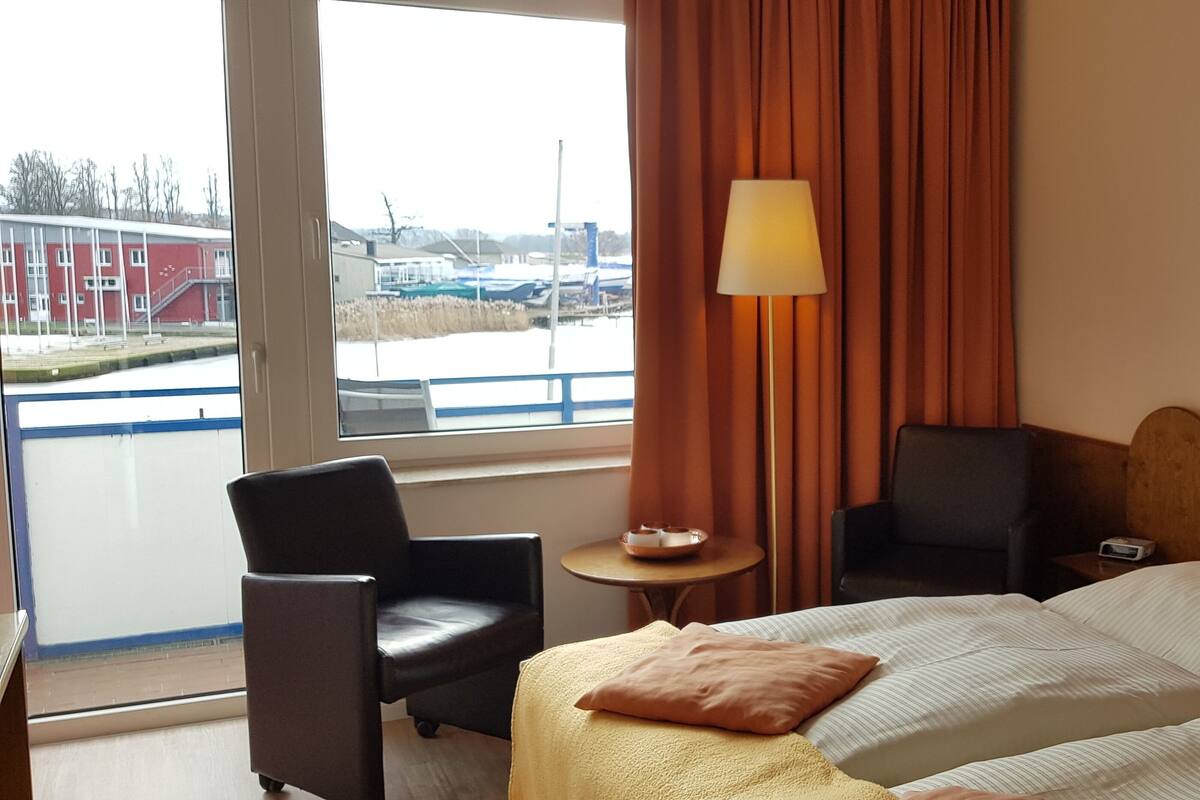 A hotel room with a bed, two chairs, a small table with a lamp, and a view of a building outside.