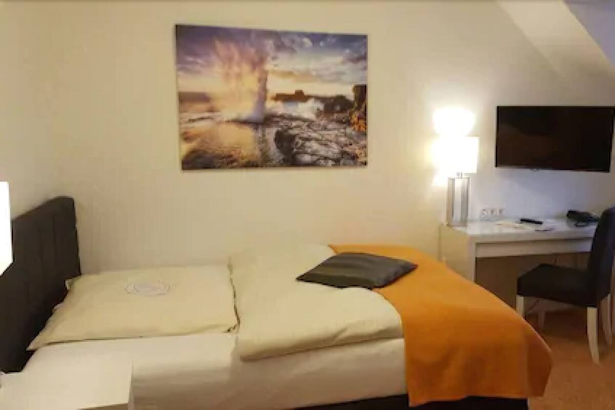 A bedroom with a bed, a desk, a chair, a TV, and a painting on the wall.