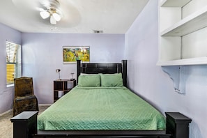 A bedroom with a bed, a chair, a nightstand, a painting, and a ceiling fan.