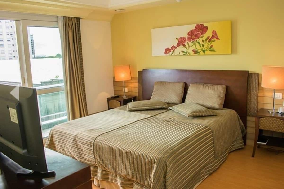 A hotel room with a large bed, two bedside tables with lamps, a flat-screen TV, and a window with curtains.