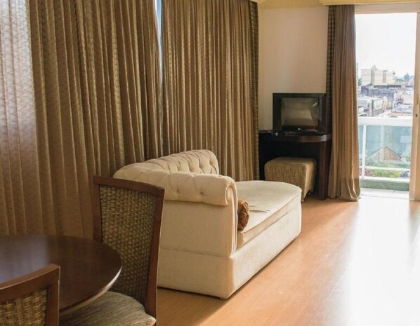Quarto de hotel com sofá, televisão, mesa pequena e vista da cidade através de amplas janelas.