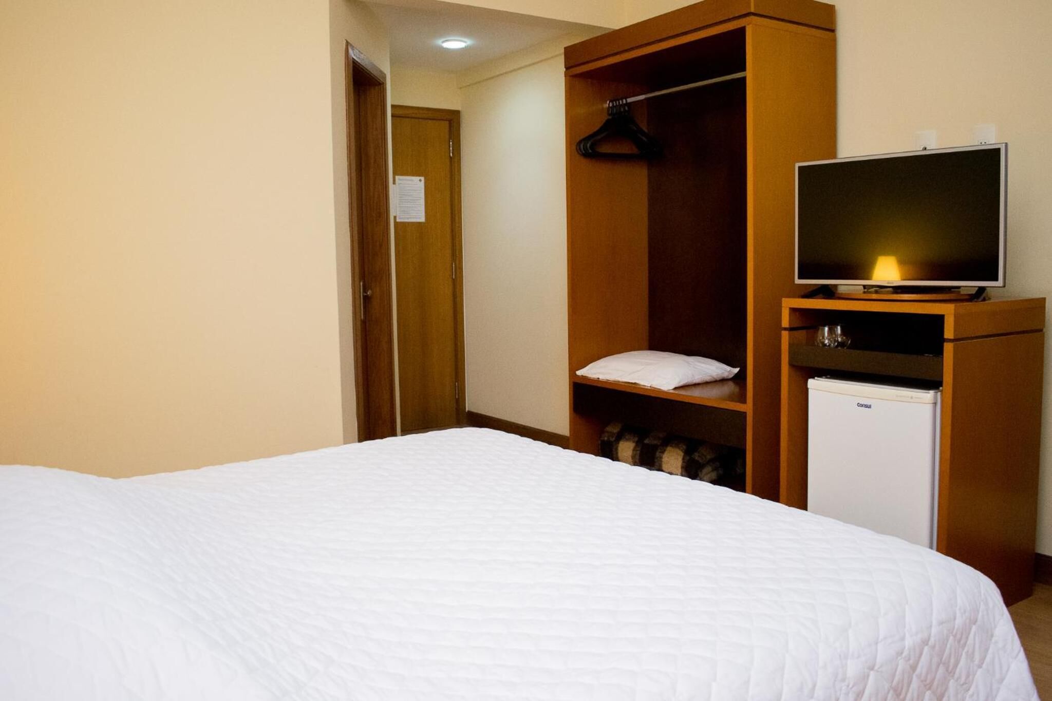A hotel room with a bed, a television, a mini-fridge, and a wooden wardrobe.