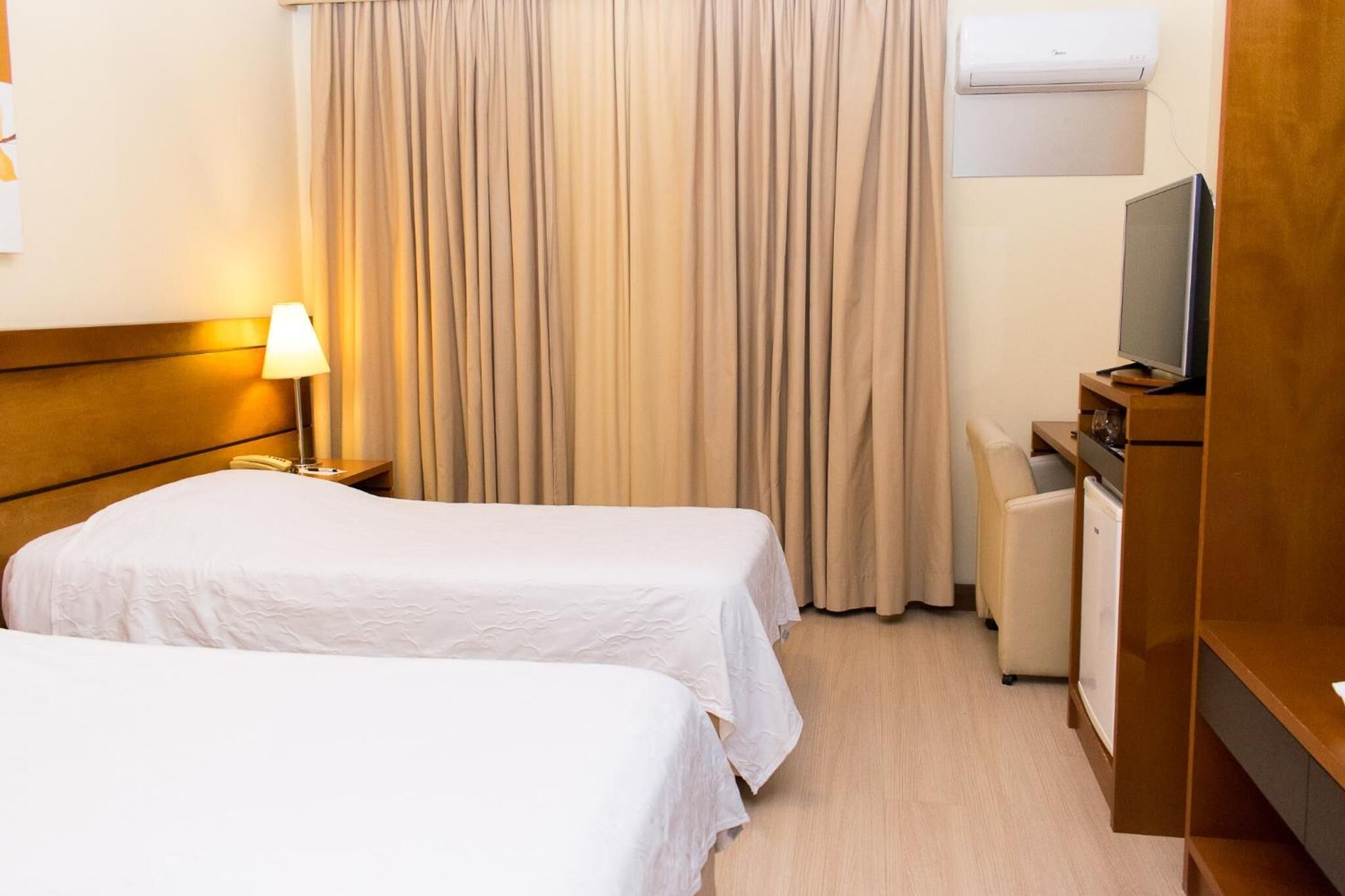 A hotel room with two beds, a desk with a computer, a lamp, a TV, and an air conditioner.