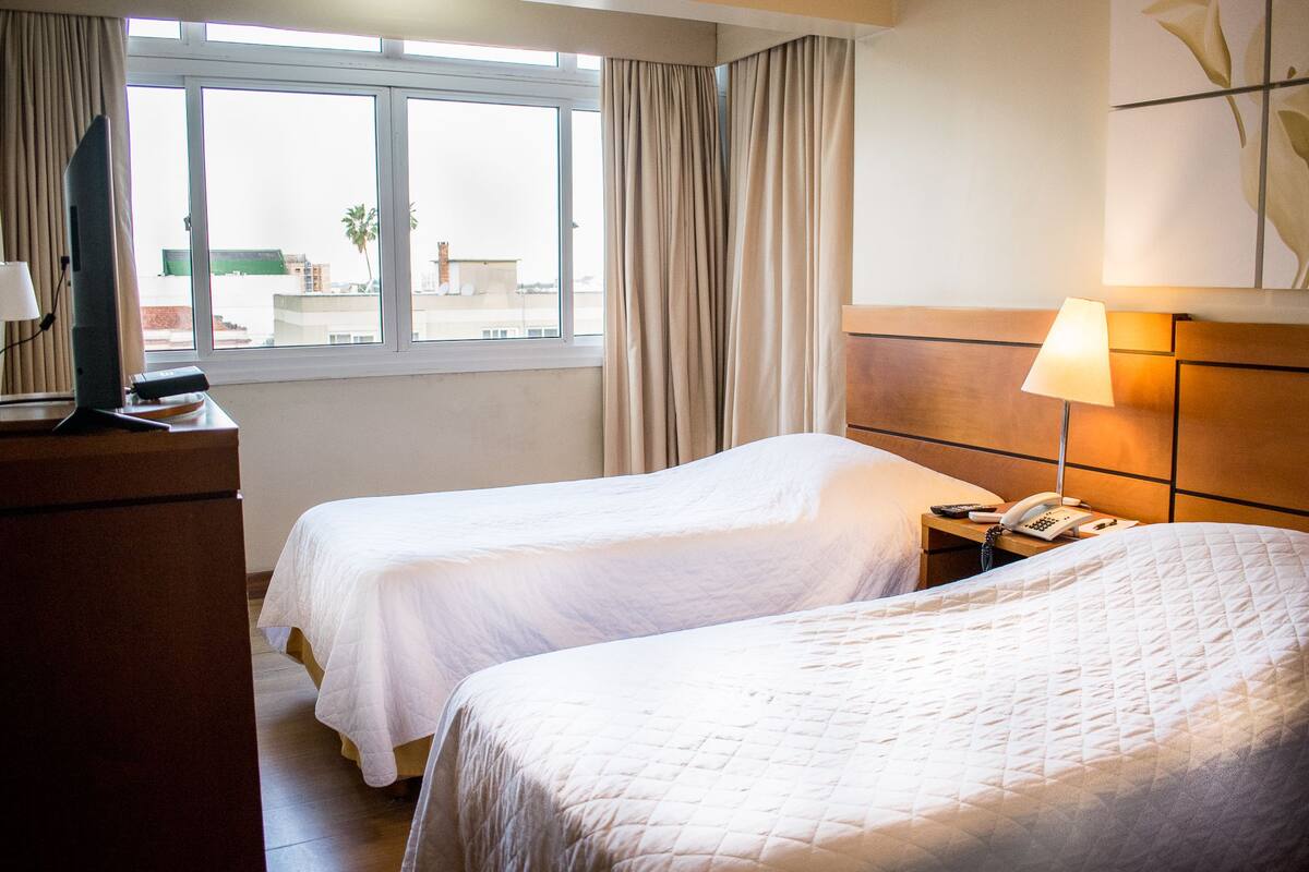 A hotel room with two beds, a TV, a bedside table with a lamp, and a window with curtains.
