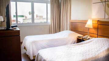 A hotel room with two beds, a TV, a bedside table with a lamp, and a window with curtains.