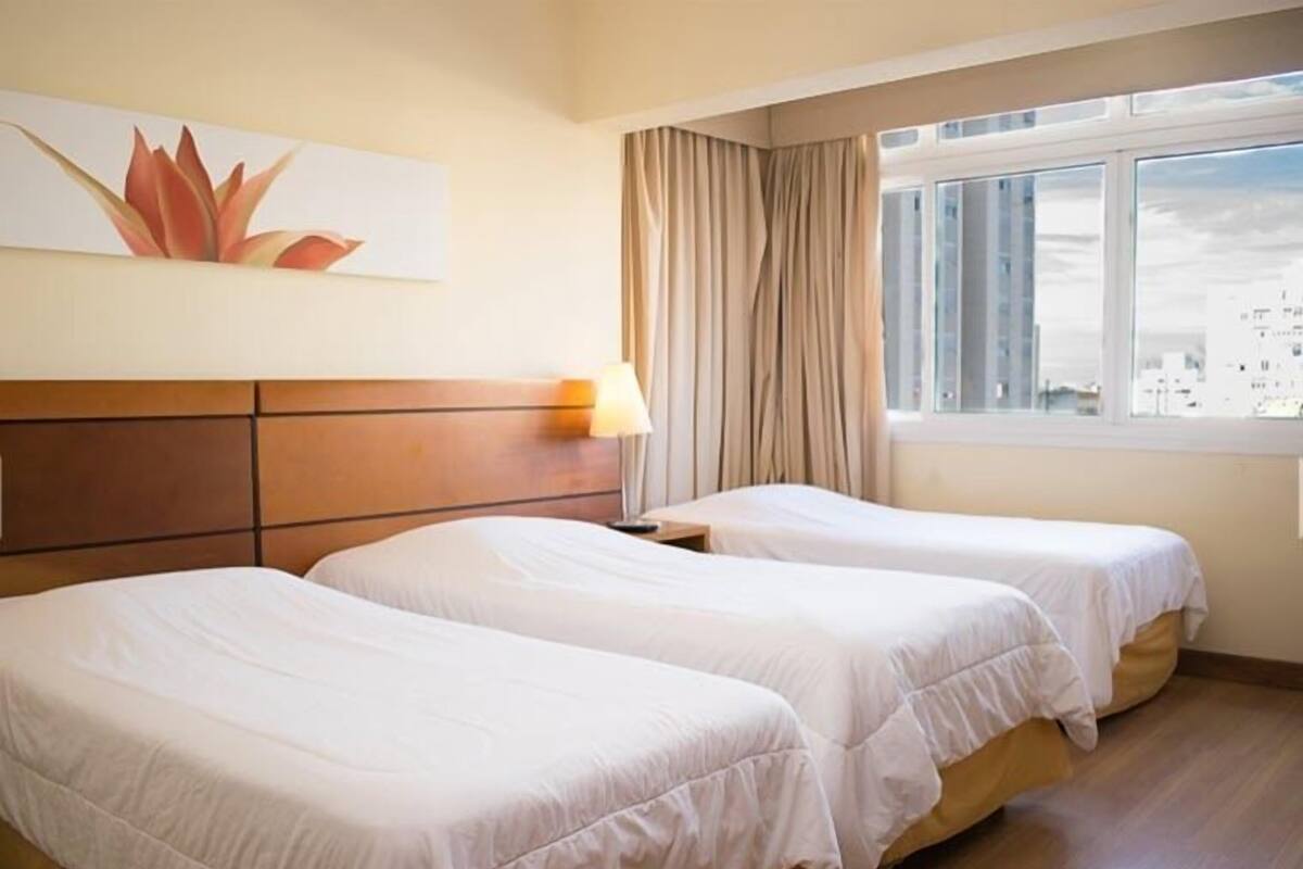 A hotel room with two single beds, a wooden headboard, a window with curtains, and a cityscape view.