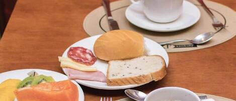 Una mesa con comida que incluye pan, queso, jamón y verduras, junto a una taza de café.
