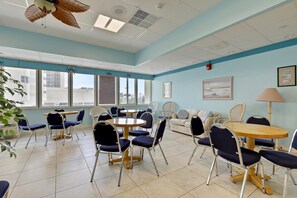 A room with multiple seating arrangements including tables and chairs, large windows, and a ceiling fan.