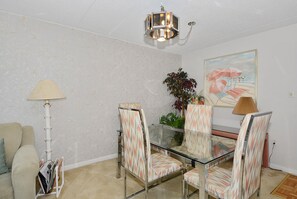 A dining area with a glass table, patterned chairs, a lamp, a plant, and a beach-themed painting on the wall. - XPLORIE ACTIVITIES & LINENS INCLUDED*! QUAY 2 BR/2 BA Ocean View Condo with Indoor/Outdoor Pools. (Ocean City)