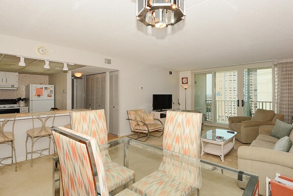 A living room with a glass coffee table, patterned chairs, a sofa, a television, and a kitchen area with a refrigerator.