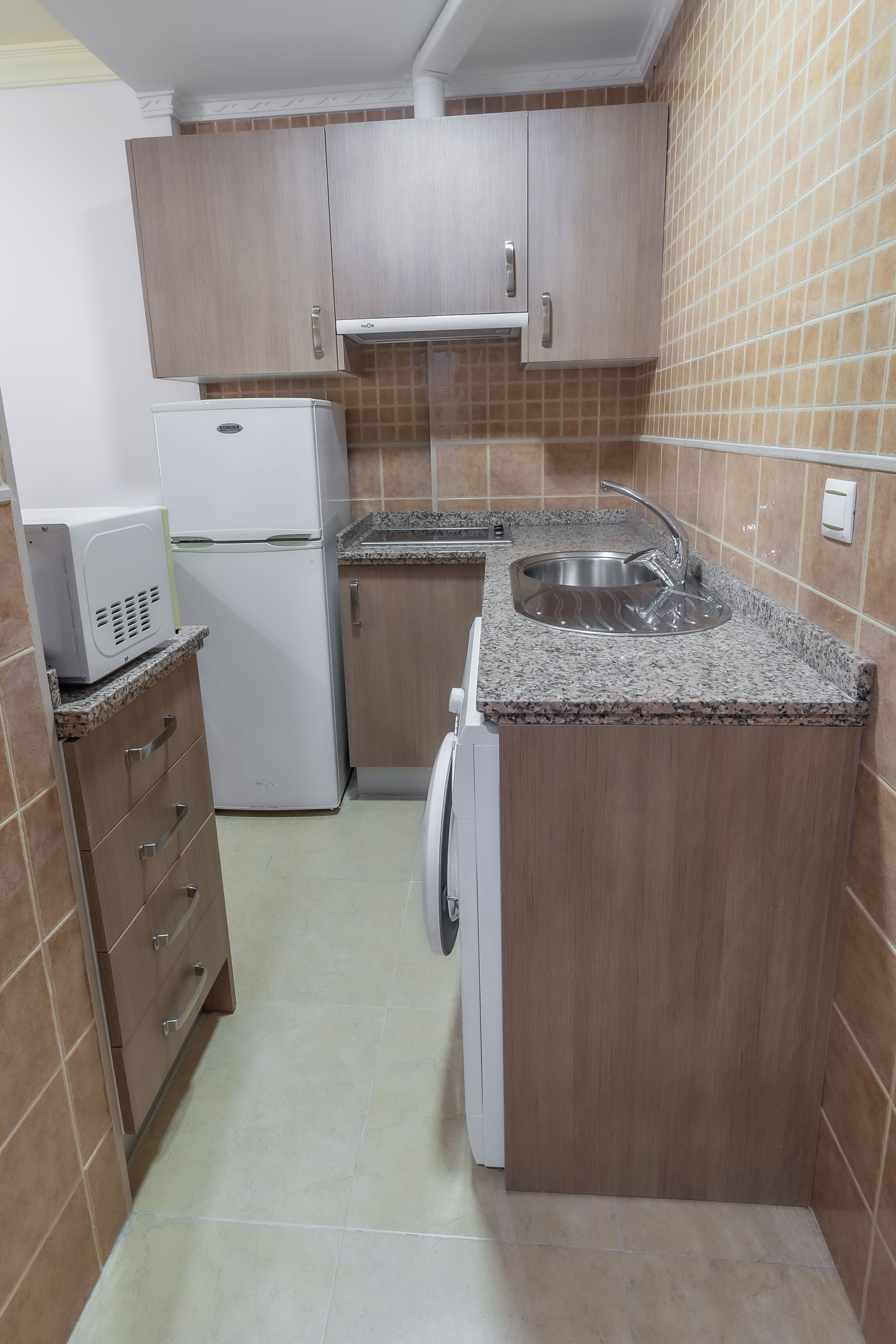 Een kleine keuken met houten kastjes, een inbouwspoelbak, een magnetron en een wasmachine.
